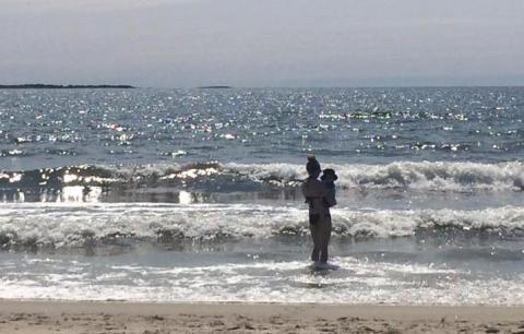 A woman holds a young child in waves at the edge of the sea.
